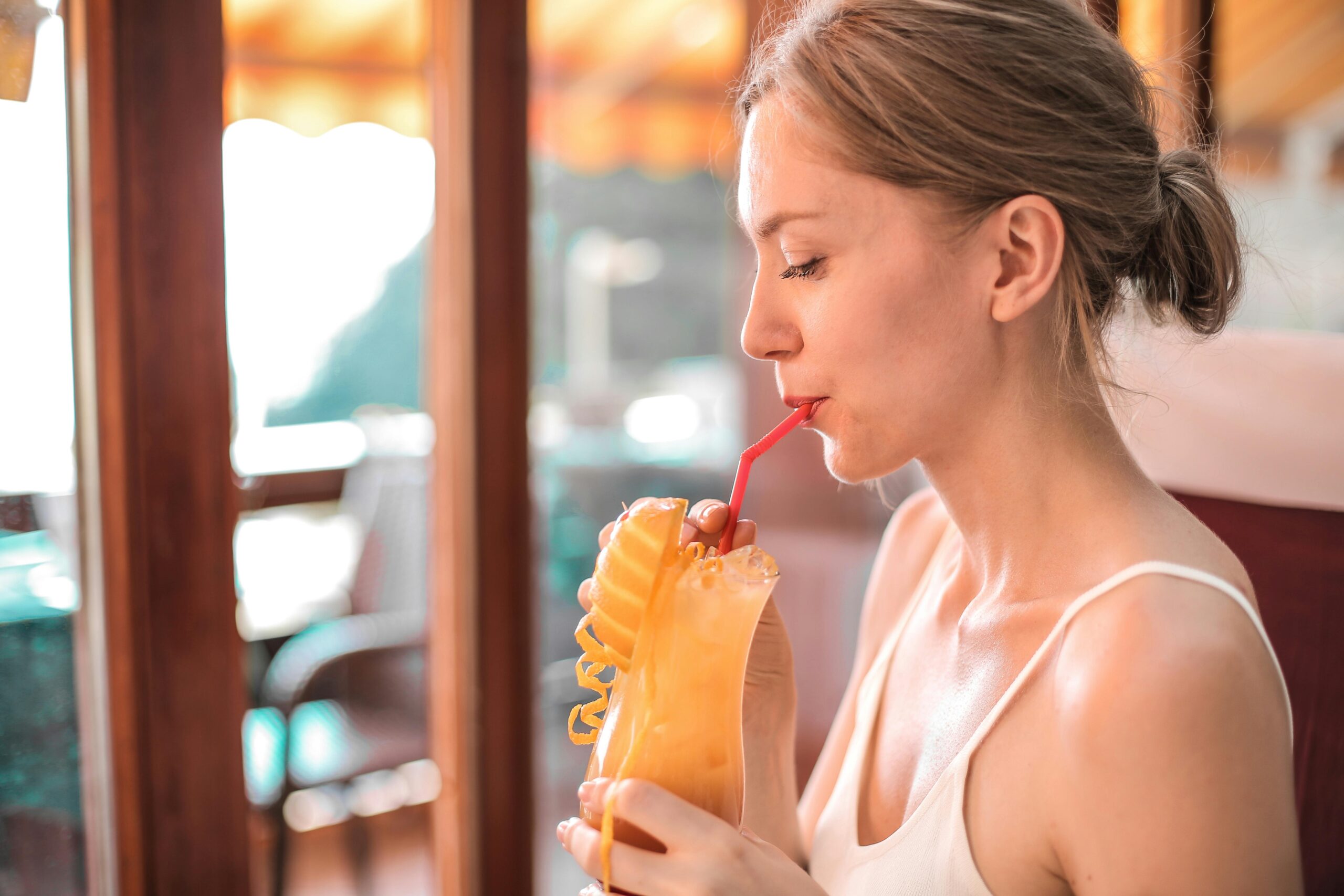 A young woman sips a refreshing citrus drink with a straw indoors, enjoying a warm and sunny day.