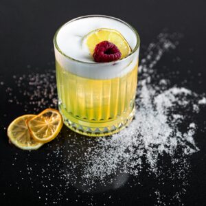A vibrant lemon cocktail garnished with raspberry, set against a black background with sugar detailing.